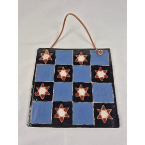 May include: A square ceramic wall hanging featuring a blue and black checkerboard pattern. Each black square contains a six-pointed star with a white center and red outline. The piece is suspended by a thin tan leather cord threaded through two holes.