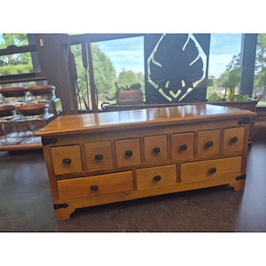 May include: A rectangular wooden storage chest featuring a natural light brown finish. The front displays a row of seven small square drawers above three wider rectangular drawers, each equipped with a small dark circular knob and black metal corner accents.