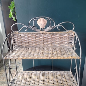 May include: A two-tiered decorative shelf with a metal frame and woven wicker shelves. The top of the shelf features a seashell design. The metal frame has a distressed, antique finish. The shelf is designed for displaying small items.