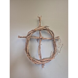 May include: A decorative wall hanging constructed from natural brown grapevine branches, featuring a circular wreath frame with a vertical and horizontal cross structure integrated into the center, showcasing raw, textured wood elements.