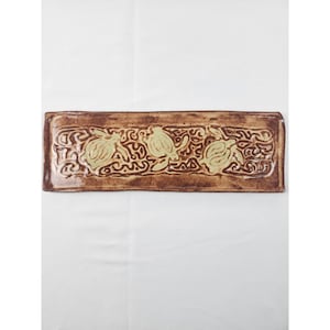 May include: A rectangular ceramic tray with a glossy brown glaze featuring three embossed light green sea turtles centered within a decorative wave-like pattern. The edges of the tray are slightly raised and have a textured, hand-crafted finish.
