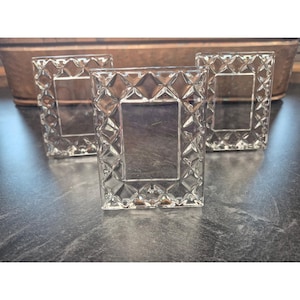 May include: Three rectangular picture frames made of clear glass, each featuring a prominent diamond-cut geometric pattern along the border. The frames are arranged in a cluster, showcasing a reflective, faceted surface texture on the outer edges.