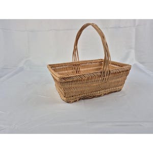 May include: A rectangular wicker basket with a tall handle. The basket is woven from light brown reeds and has a divided interior. The handle arches over the top, providing a sturdy grip. The basket sits on a white surface.