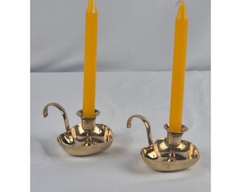 May include: Two polished brass candle holders, each featuring a curved handle and a wide, shallow base. A single bright yellow taper candle is inserted into the central socket of each holder, standing upright against a plain white background.