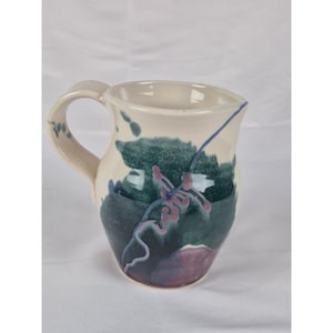 May include: Cream-colored ceramic pitcher with a rounded handle and a spout. The pitcher is decorated with abstract green, blue, and purple brushstrokes. A thin blue line and pink accents add detail to the design. The pitcher is suitable for serving beverages.