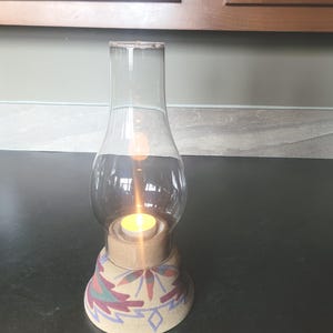 May include: A clear glass hurricane candle holder with a lit candle inside. The base is beige with a colorful pattern of leaves and geometric shapes in red, green, and purple. The candle holder is on a dark surface.