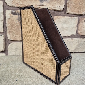 May include: A decorative magazine holder with a woven beige front and sides, framed with dark brown leather trim. The interior is a dark brown suede-like material. The edges are stitched with a light-colored thread.