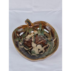 May include: A ceramic apple-shaped bowl with a stem. The bowl features a glossy glaze with shades of brown, green, and cream. The interior has a mottled pattern, and the rim is a darker brown. This decorative bowl is suitable for serving or display.