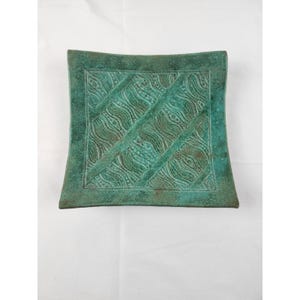 May include: A square ceramic dish with a textured, teal-green patina finish. The center features a raised, repeating pattern of diagonal bands filled with intricate, wavy lines and small dots, framed by a smooth, flat border edge.