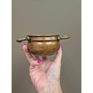 May include: A small, round copper pot with a smooth, metallic finish and a warm, reddish-brown hue. The vessel features two ornate, curved brass handles attached to opposite sides of the rim, providing a distinct contrast in color and texture.