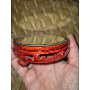 May include: A round, reddish-brown wooden display stand featuring an openwork carved design along the sides and three small feet. The top surface is flat and circular, providing a stable base for objects, with a smooth, polished finish.