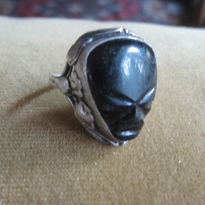 May include: A silver ring with a black carved stone in the shape of a face. The stone has a detailed design and is set in a silver band.