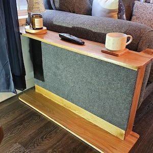 May include: A wooden sofa arm table with a gray fabric panel and a lower shelf. The table has a natural wood finish and is designed to fit over the arm of a sofa. A remote control and a mug are on the table.