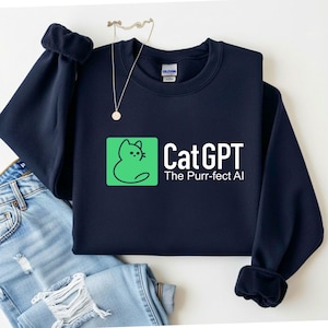 Op de afbeelding: Marineblauwe sweatshirt met de tekst "CatGPT" en de slogan "The Purr-fect AI". Het ontwerp bevat een groene vierkant met een kattenillustratie. De sweatshirt wordt getoond met een blauwe jeans en een ketting.