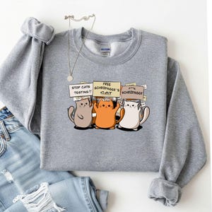 May include: A gray sweatshirt featuring a graphic of three cartoon cats holding signs that read "STOP CATS TESTING!!!", "FREE SCHRODINGER'S CAT", and "F**K SCHRODINGER". The cats are in shades of brown, orange, and white.