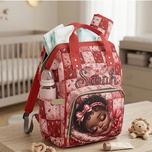 May include: Red and pink quilted baby backpack with the name "Sarah" embroidered on the front. The backpack has a side pocket with a baby bottle, and a front pocket with a sleeping baby illustration. Diapers and a changing pad are visible.