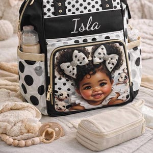 Peut inclure: Sac à langer noir et blanc à pois avec le nom "Isla". La poche avant présente une fillette avec des nœuds. Le sac a des poches, une poignée et un porte-biberon. Une tétine, un petit sac et une couverture sont également visibles.