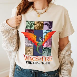 May include: Beige t-shirt with a dragon graphic, featuring a grid of dragon illustrations and a central dragon with orange and red wings. The text "Wings of Fire: The Eras Tour" is displayed in red and white.