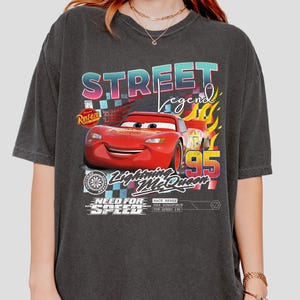 May include: Dark gray t-shirt featuring a graphic of Lightning McQueen from the movie Cars. The design includes the text "Street Legend", "Lightning McQueen 95", and "Need for Speed". The shirt has a vintage, distressed look.