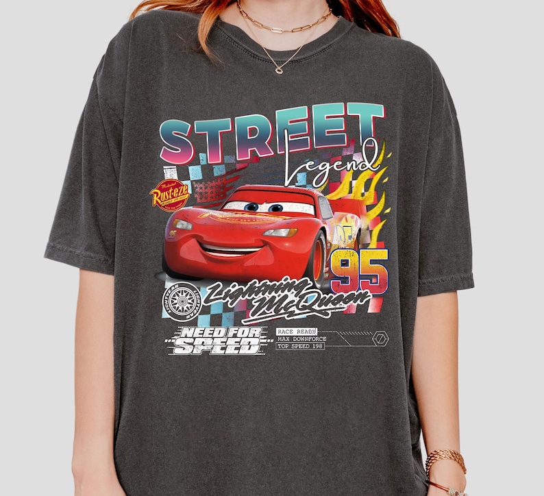 May include: Dark gray t-shirt featuring a graphic of Lightning McQueen from the movie Cars. The design includes the text "Street Legend", "Lightning McQueen 95", and "Need for Speed". The shirt has a vintage, distressed look.