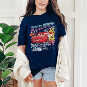May include: Navy blue t-shirt featuring a graphic of Lightning McQueen from the movie Cars. The design includes the text "Street Legend" and "Lightning McQueen 95" with racing-themed elements. The shirt is worn by a person.