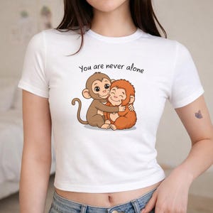May include: White t-shirt with a cartoon image of two monkeys embracing. One monkey is brown, the other is orange. The text "You are never alone" is above the monkeys in black script.