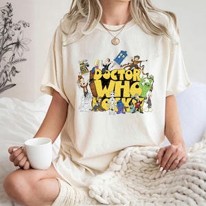 May include: Cream-colored t-shirt featuring a colorful graphic of characters and the words "Doctor Who" in yellow. The design includes various figures from the show. The person is holding a white mug.
