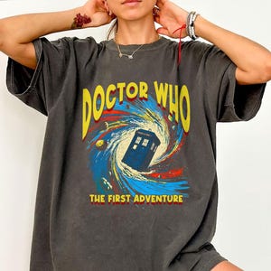 May include: Dark gray t-shirt featuring a colorful graphic from the Doctor Who series. The design includes the text "DOCTOR WHO" in yellow, a blue police box, and the phrase "THE FIRST ADVENTURE". The shirt has a vintage, worn look.
