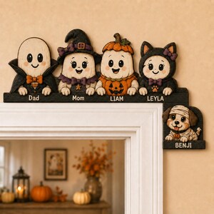 May include: A Halloween-themed wooden door corner sign featuring five cartoon characters: a ghost labelled Dad, a witch ghost labelled Mom, a pumpkin ghost labelled LIAM, a cat ghost labelled LEYLA, and a mummy puppy labelled BENJI on the side.