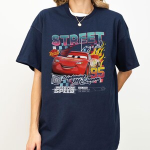May include: Navy blue t-shirt featuring a graphic of Lightning McQueen from the movie Cars. The design includes the text "STREET LEGEND", "Lightning McQueen 95", and "NEED FOR SPEED". The graphic has a colorful, retro style.