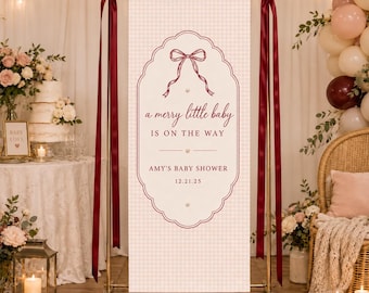 May include: A vertical rectangular banner with a light pink gingham pattern, featuring a dark red decorative border, a bow illustration, and the text: a merry little baby IS ON THE WAY, AMY'S BABY SHOWER, 12.21.25. It hangs from a gold metal stand.