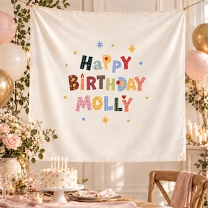 May include: A cream-colored fabric wall hanging featuring the text "HAPPY BIRTHDAY MOLLY" in colorful, patterned block letters. The design includes small star and dot accents in blue, yellow, and green, creating a festive display for a celebration.