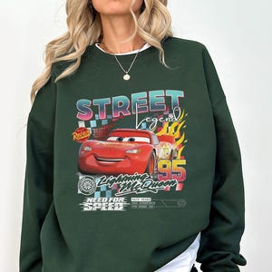 May include: Dark green sweatshirt featuring a graphic of Lightning McQueen from the movie Cars. The design includes the text "Street Legend", "Lightning McQueen", and "Need for Speed". The number "95" is also prominently displayed.