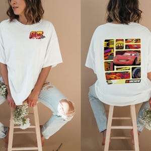 May include: White t-shirt featuring a colorful graphic of a race car on the back and a smaller design on the front. The back graphic includes the text "NACHOW". The shirt is worn with ripped blue jeans.