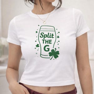 Pode incluir: Top curto branco com um gráfico verde de um copo de cerveja e o texto "Split the G". O design inclui trevos e estrelas, perfeito para as celebrações do Dia de São Patrício.