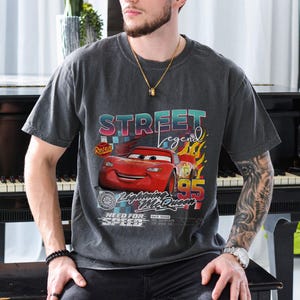 May include: Dark gray t-shirt featuring a graphic of Lightning McQueen from the movie Cars. The design includes the text "Street Legend" and "95" with flames. The shirt has a vintage, worn look.
