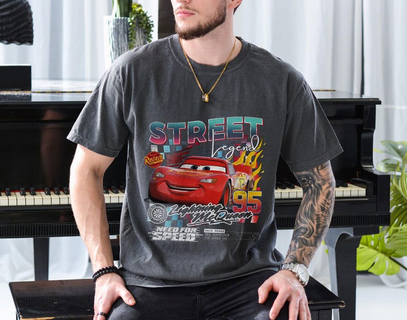 May include: Dark gray t-shirt featuring a graphic of Lightning McQueen from the movie Cars. The design includes the text "Street Legend" and "95" with flames. The shirt has a vintage, worn look.