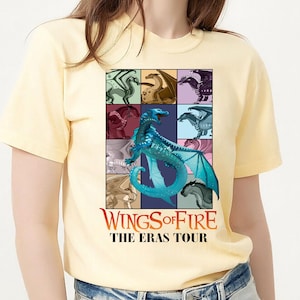 May include: A light yellow t-shirt featuring a colorful graphic of dragons in a grid pattern. The graphic includes the text "Wings of Fire The Eras Tour" in red and white. The shirt is made of a soft material.