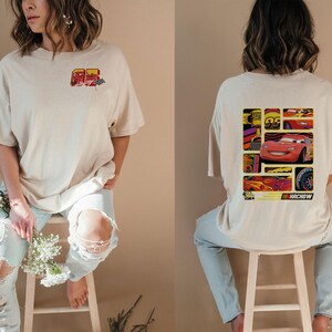 May include: Beige t-shirt featuring a graphic of a red race car with the number 95. The back of the shirt displays a collage of images related to the car, including the text "KA-CHOW".