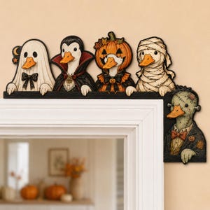 May include: A decorative door corner sign featuring five cartoon ducks dressed in Halloween costumes: a ghost, a vampire, a pumpkin, a mummy, and a Frankenstein monster. The ducks are arranged along a black L-shaped frame with orange beaks.