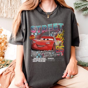 May include: Dark gray t-shirt featuring a graphic of Lightning McQueen from the movie Cars. The design includes the text "STREET LEGEND" in a colorful font, the number "95", and the phrase "NEED FOR SPEED".