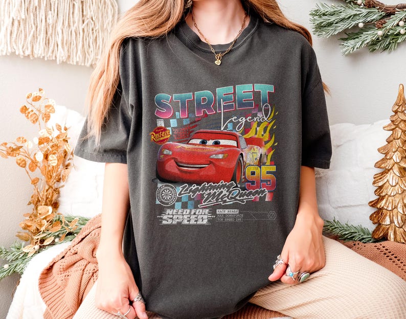 May include: Dark gray t-shirt featuring a graphic of Lightning McQueen from the movie Cars. The design includes the text "STREET LEGEND" in a colorful font, the number "95", and the phrase "NEED FOR SPEED".