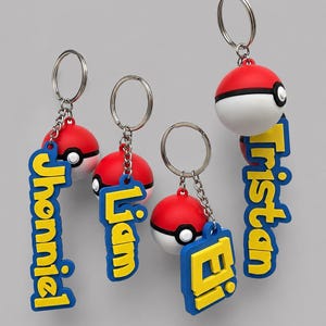 May include: Four keychains featuring spherical red and white ball charms attached to silver metal rings and chains. Each keychain includes a blue rectangular tag with yellow lettering displaying the names Jhonniel, Liam, Ei, and Tristan.