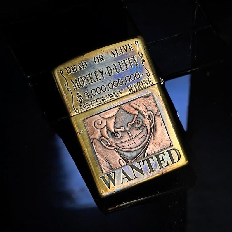 May include: A brass-colored lighter featuring a detailed engraving of a smiling cartoon character. The lighter has the text "DEAD OR ALIVE MONKEY-D-LUFFY $3,000,000,000- MARINE WANTED" etched on its surface. The design is intricate and eye-catching.