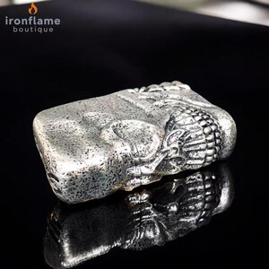 May include: A silver rectangular lighter with an embossed design. The lighter has a textured surface and a decorative element on one end. The logo "ironflame boutique" is in the upper left corner.