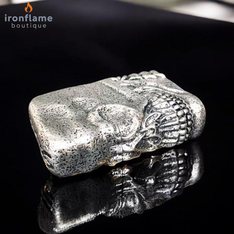 May include: A silver rectangular lighter with an embossed design. The lighter has a textured surface and a decorative element on one end. The logo "ironflame boutique" is in the upper left corner.