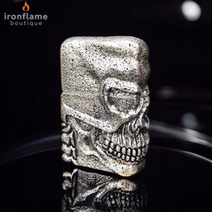 May include: A silver-coloured lighter with a skull design. The lighter has a textured surface and a hinged lid. The skull's features, including the eye sockets, nose, and teeth, are clearly defined. The lighter is displayed on a reflective black surface.