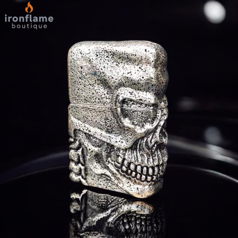 May include: A silver-coloured lighter with a skull design. The lighter has a textured surface and a hinged lid. The skull's features, including the eye sockets, nose, and teeth, are clearly defined. The lighter is displayed on a reflective black surface.