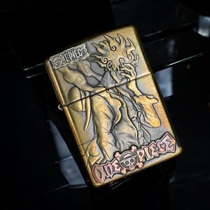 May include: A brass-toned metal lighter featuring a detailed relief of a character from the anime series "One Piece." The design includes the text "ONE PIECE" and a skull and crossbones emblem. The lighter has a hinged lid and a brushed finish.