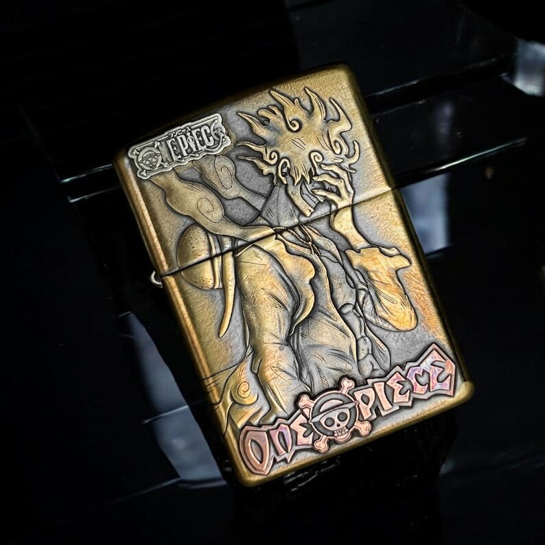 May include: A brass-toned metal lighter featuring a detailed relief of a character from the anime series "One Piece." The design includes the text "ONE PIECE" and a skull and crossbones emblem. The lighter has a hinged lid and a brushed finish.